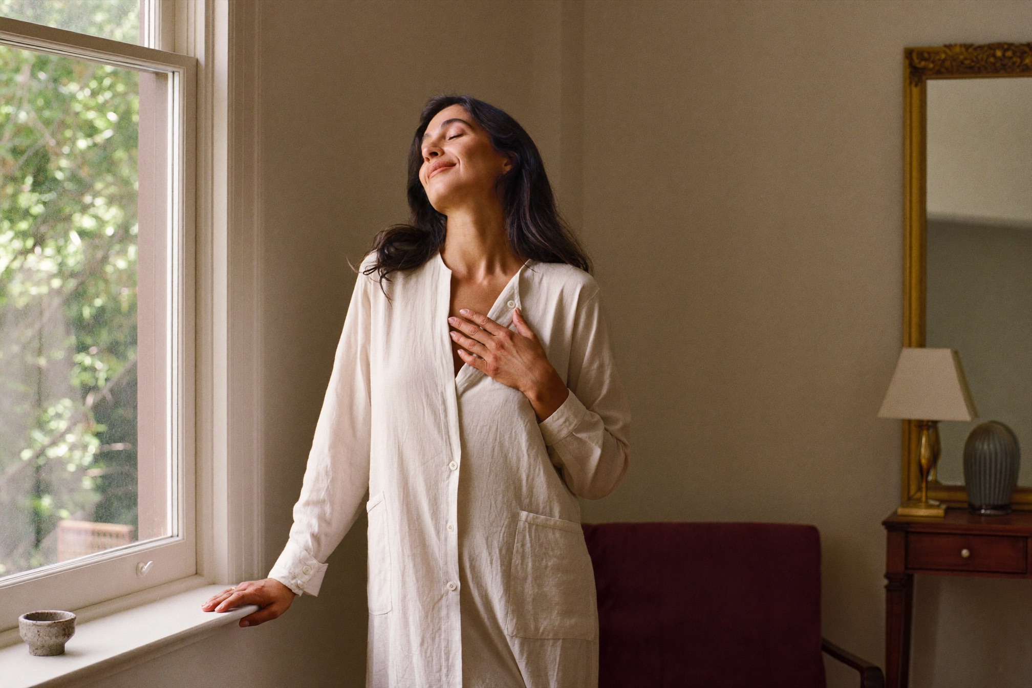 A mother exhaling at ease by a sunlit window, one hand resting gently over her heart